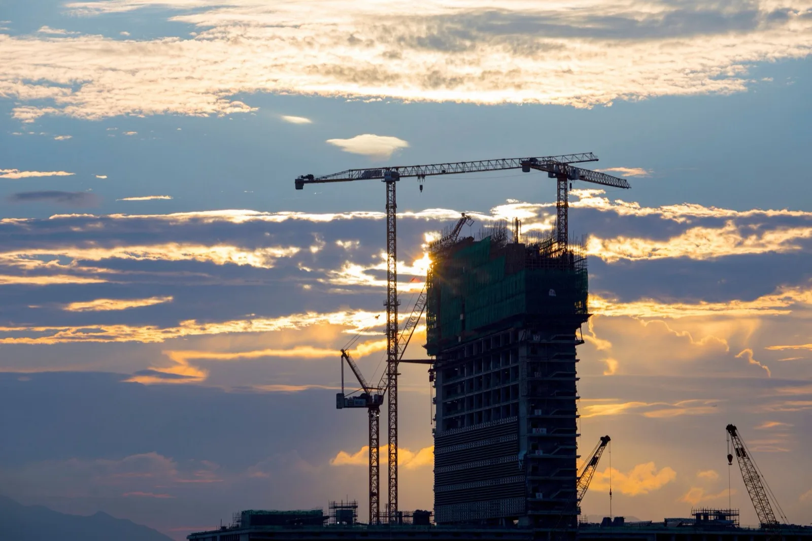 Tower under construction with two cranes in silhouette at sunset
