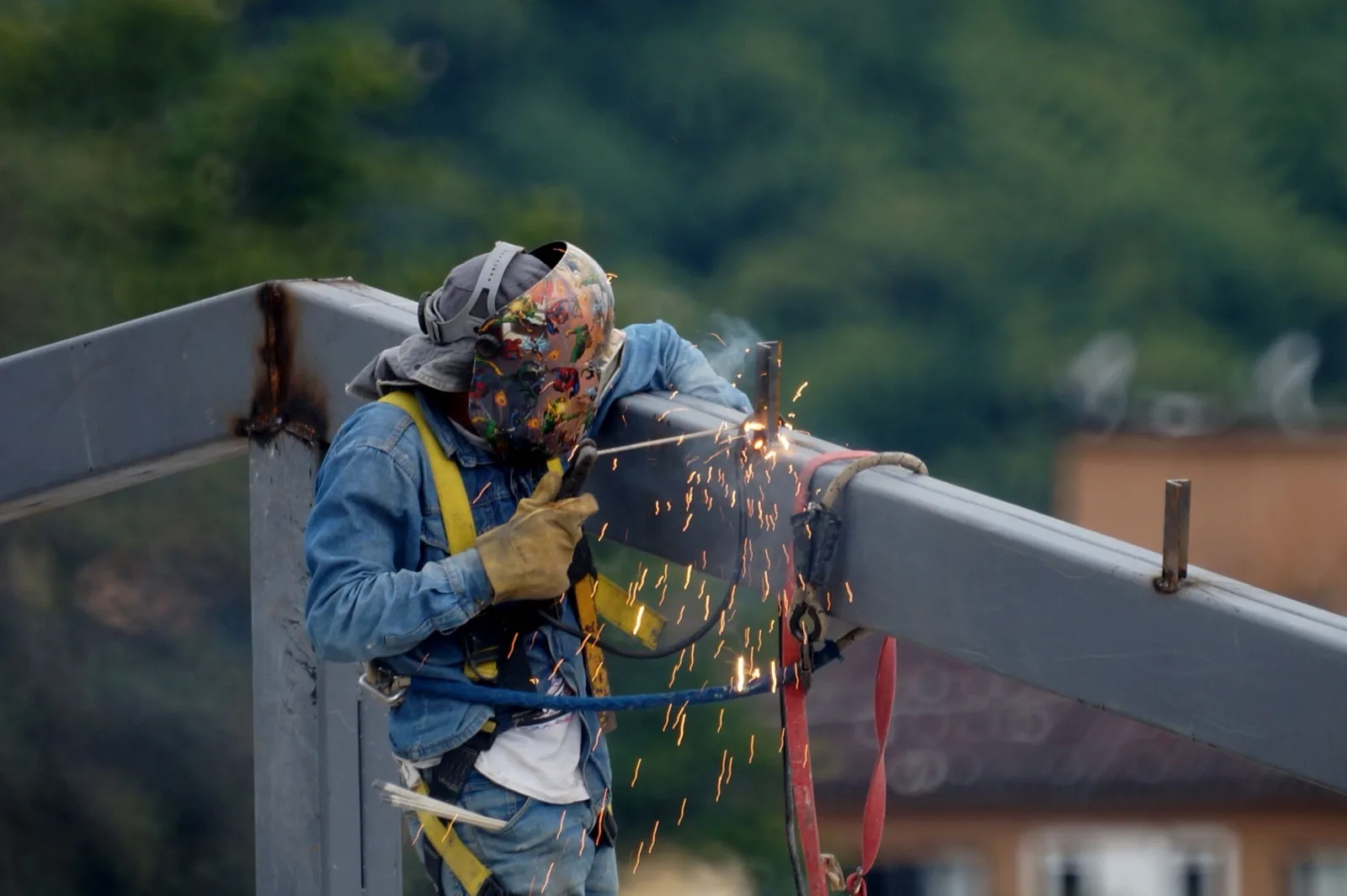 Ironworker in a welding mask on a steel beam, sparks flying, trees behind