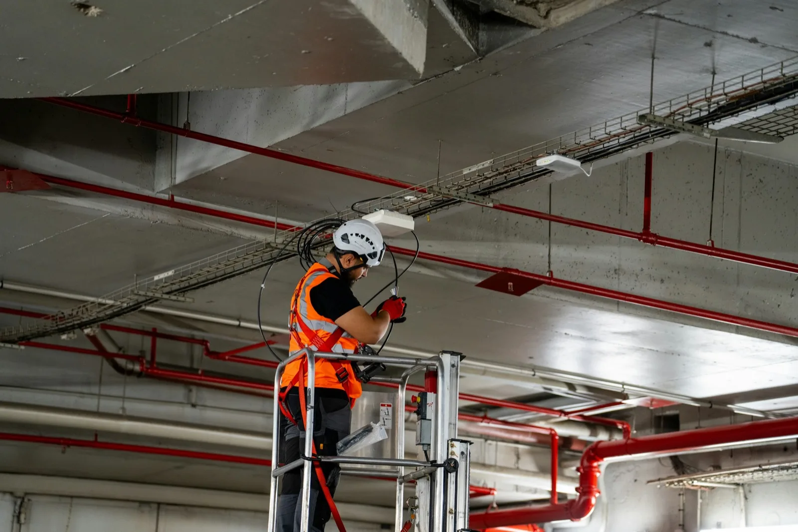 Technician on a lift installing cabling under a ceiling of trays and sprinkler pipe