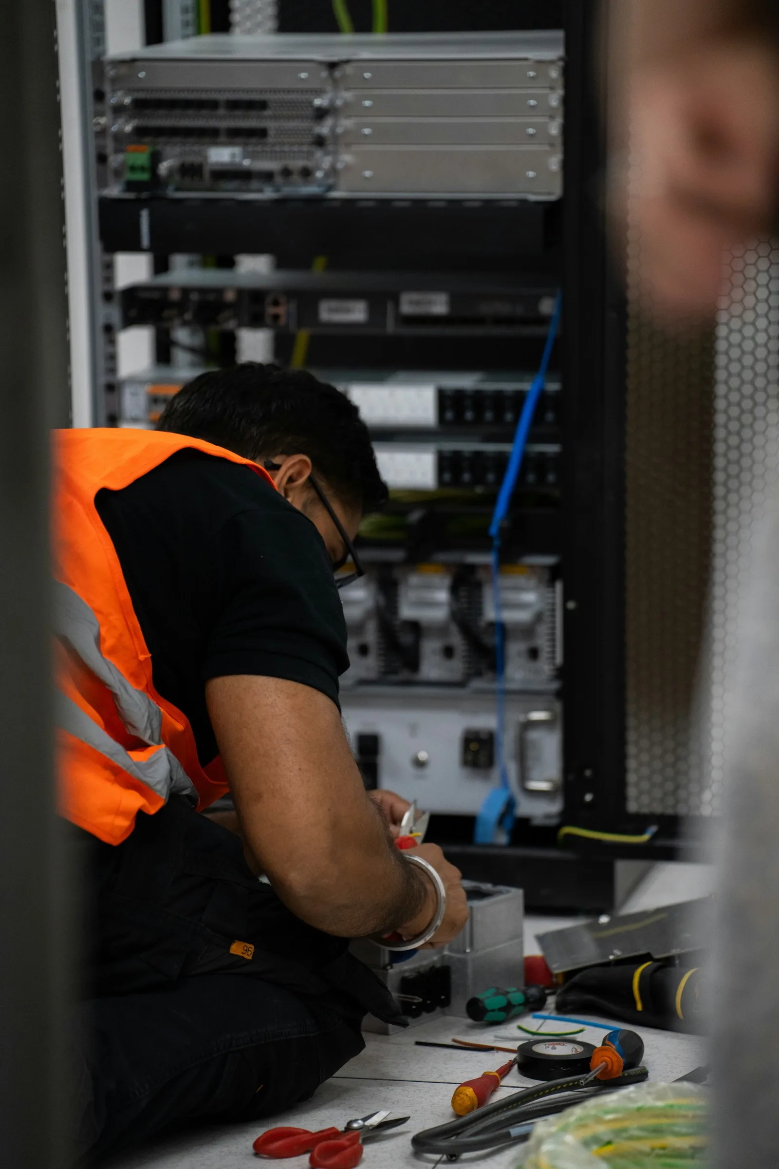 Technician in an orange vest kneeling at an open network rack with a tester